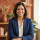 A friendly South Asian woman smiling for a professional portrait in a warm, terracotta-toned setting.